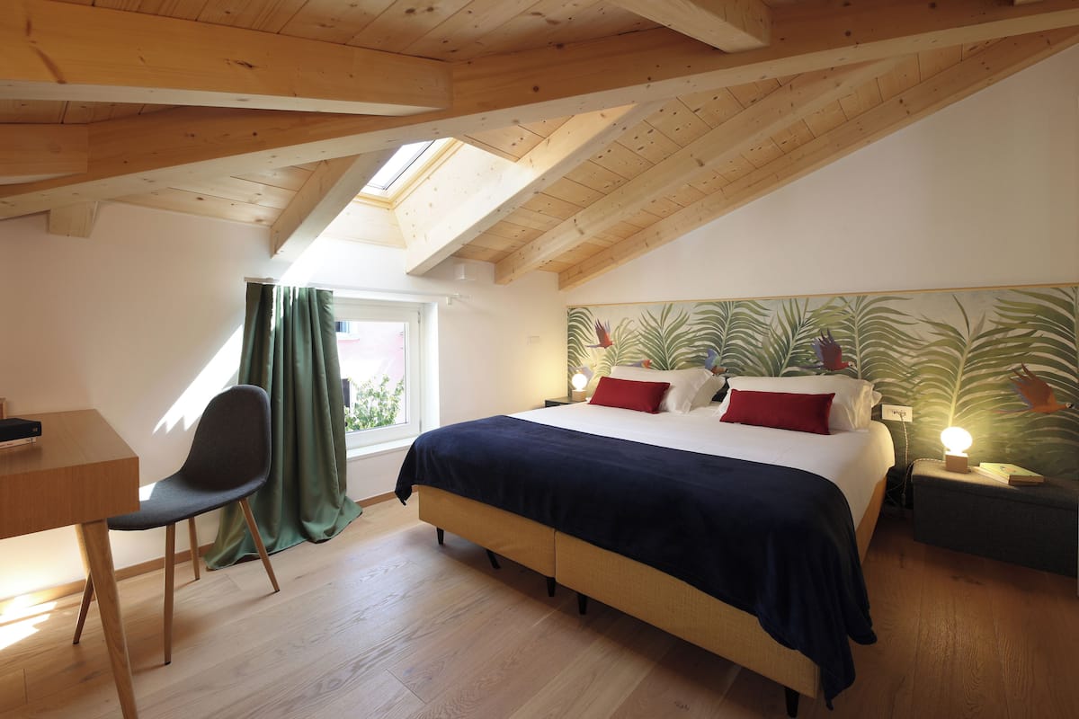 A bedroom with a sloped ceiling, a bed with a dark blue bedspread, a desk with a chair, and a window with curtains.