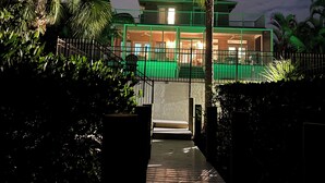 A modern two-story building with a green facade, a wooden deck leading to the entrance, and a palm tree in the foreground. - Siesta Key Complete House - Pool - Jacuzzi - 3 Bedroom - 2 Bath - Screened Patio (Sarasota)