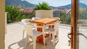 A balcony with a wooden table and chairs, a view of mountains, and a tiled floor.