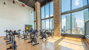 Une salle de sport avec des vélos d’appartement disposés en rangées, de grandes fenêtres donnant sur la ville et un mur en briques.