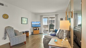 A living room with a sofa, armchair, TV, wooden table, and a view of the sea through a sliding door.
