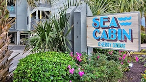 A wooden sign for Sea Cabin in Cherry Grove, surrounded by lush greenery and a building in the background.