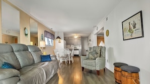 A spacious living area with a leather sofa, patterned armchair, and wooden stools. A dining table with chairs is visible in the background.