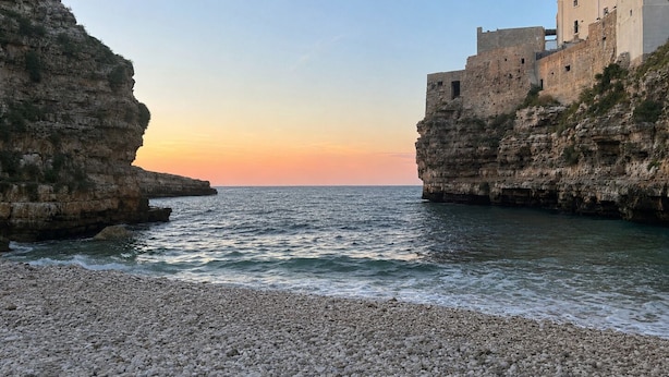 A coastal scene with rocky cliffs, a pebbled beach, and a building with a flat roof.