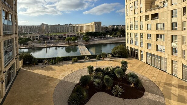 Un patio central con zonas ajardinadas, rodeado de edificios modernos, una masa de agua y una vista distante del paisaje urbano.