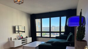 Una sala de estar moderna con un ventanal grande, un televisor de pantalla plana, una mesa de centro blanca y un sofá de color oscuro.