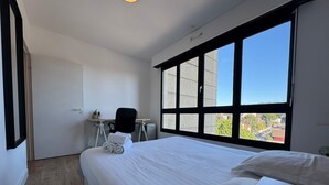 Habitación de hotel con una cama grande, un escritorio con una silla y vistas a la ciudad a través de ventanas amplias.