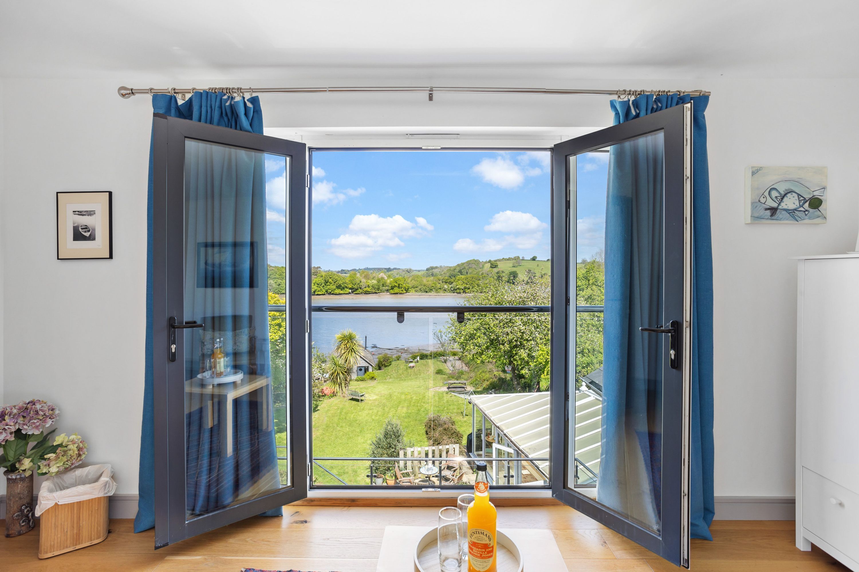A room with an open sliding glass door leading to a balcony with a view of a lake and greenery.