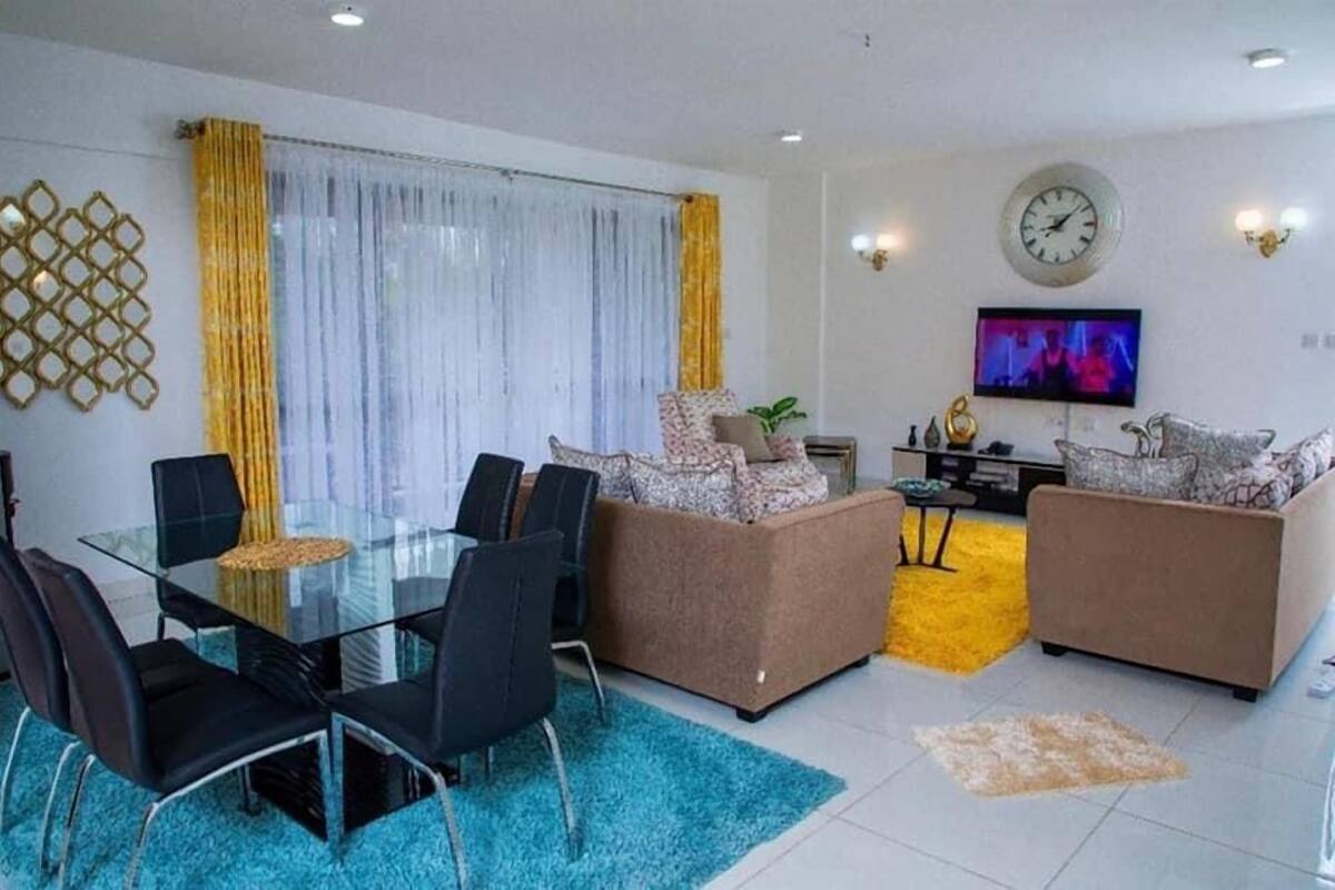 A modern living room with a glass dining table, two sofas, a TV, and a decorative wall piece.