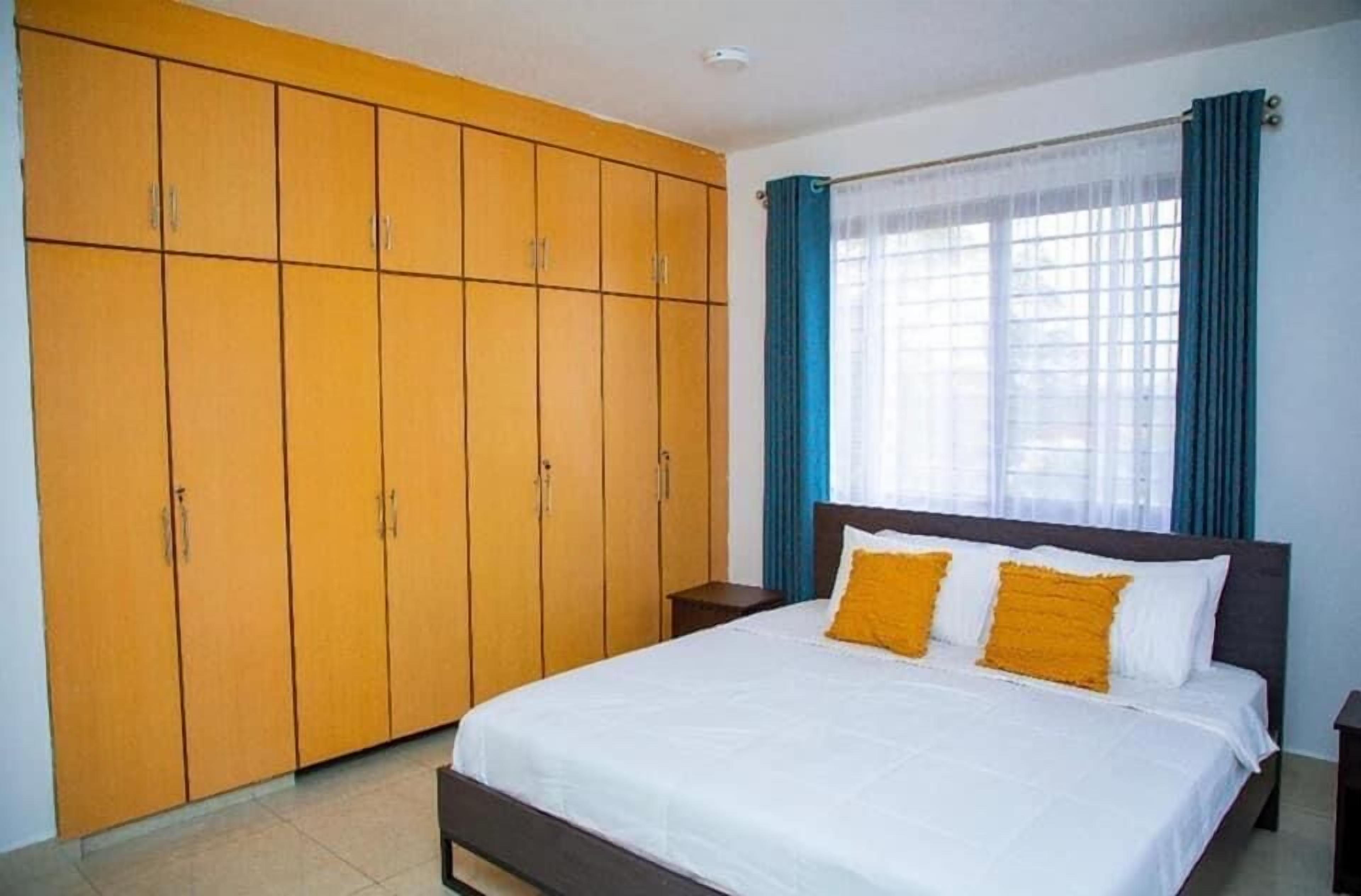 A bedroom with a large wardrobe, a bed with white bedding and two yellow pillows, and a window with blue curtains.