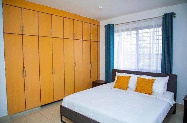 A bedroom with a large wardrobe, a bed with white bedding and two yellow pillows, and a window with blue curtains.