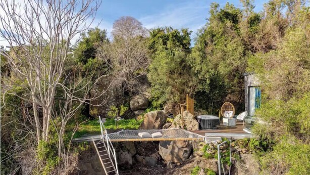 A natural setting with a wooden bridge, a ladder, and a seating area with chairs and a table.