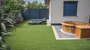 Un jardin avec une pelouse artificielle, deux chaises longues, un bain à remous en bois et une porte de garage bleue.
