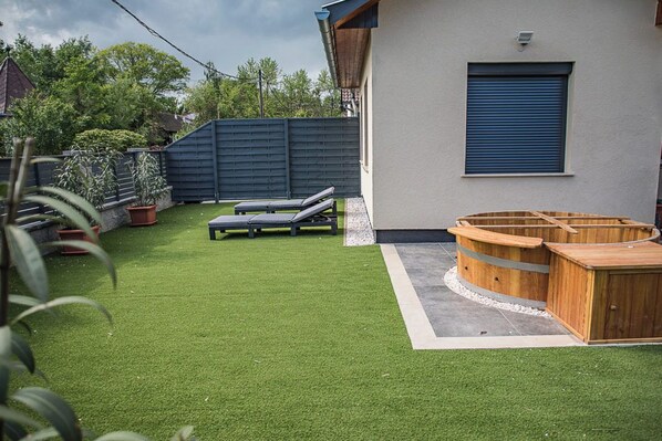 Un jardín con césped artificial, dos sillones de descanso, una bañera de hidromasaje de madera y una puerta de garaje azul.