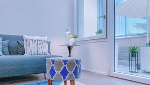 Un salon moderne avec un canapé, un pouf à motifs, une petite table avec une plante et une vue dégagée sur l’extérieur grâce à une grande fenêtre.