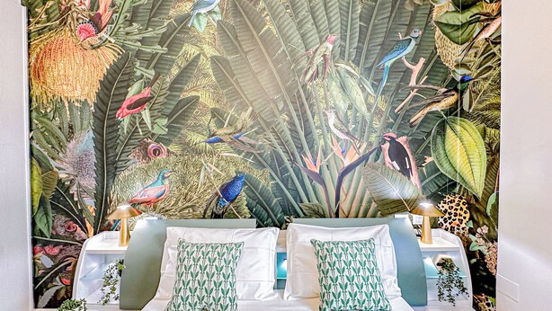 A bedroom with a large mural of birds and plants on the wall, a bed with white linens, and two pillows with a leaf pattern.