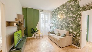 A living room with a herringbone floor, a sofa, a TV, a plant, and a wall with a tropical leaf pattern.