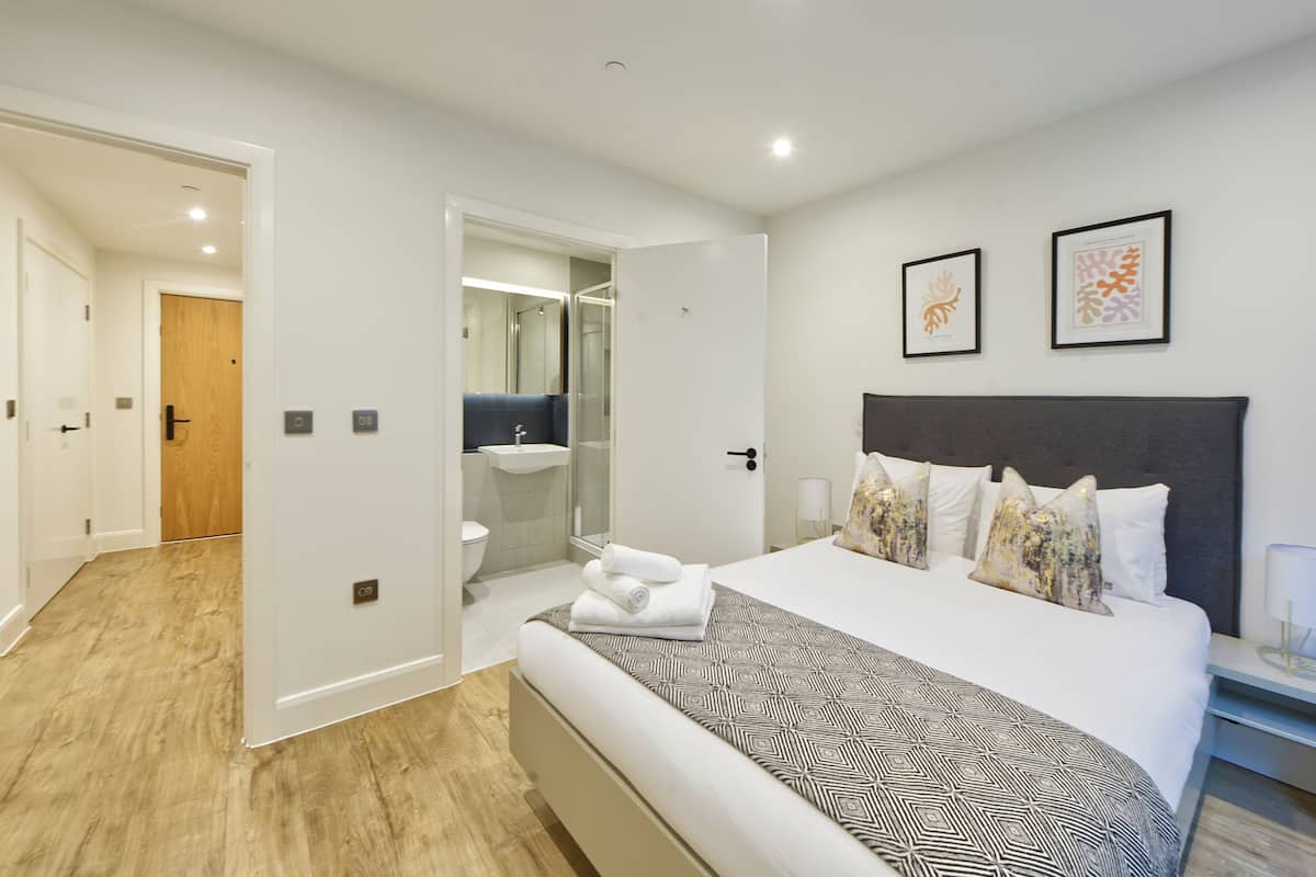 Une chambre d’hôtel moderne dotée d’un grand lit, d’une table de chevet, d’une salle de bain avec une douche en verre, et de deux œuvres d’art encadrées accrochées au mur.