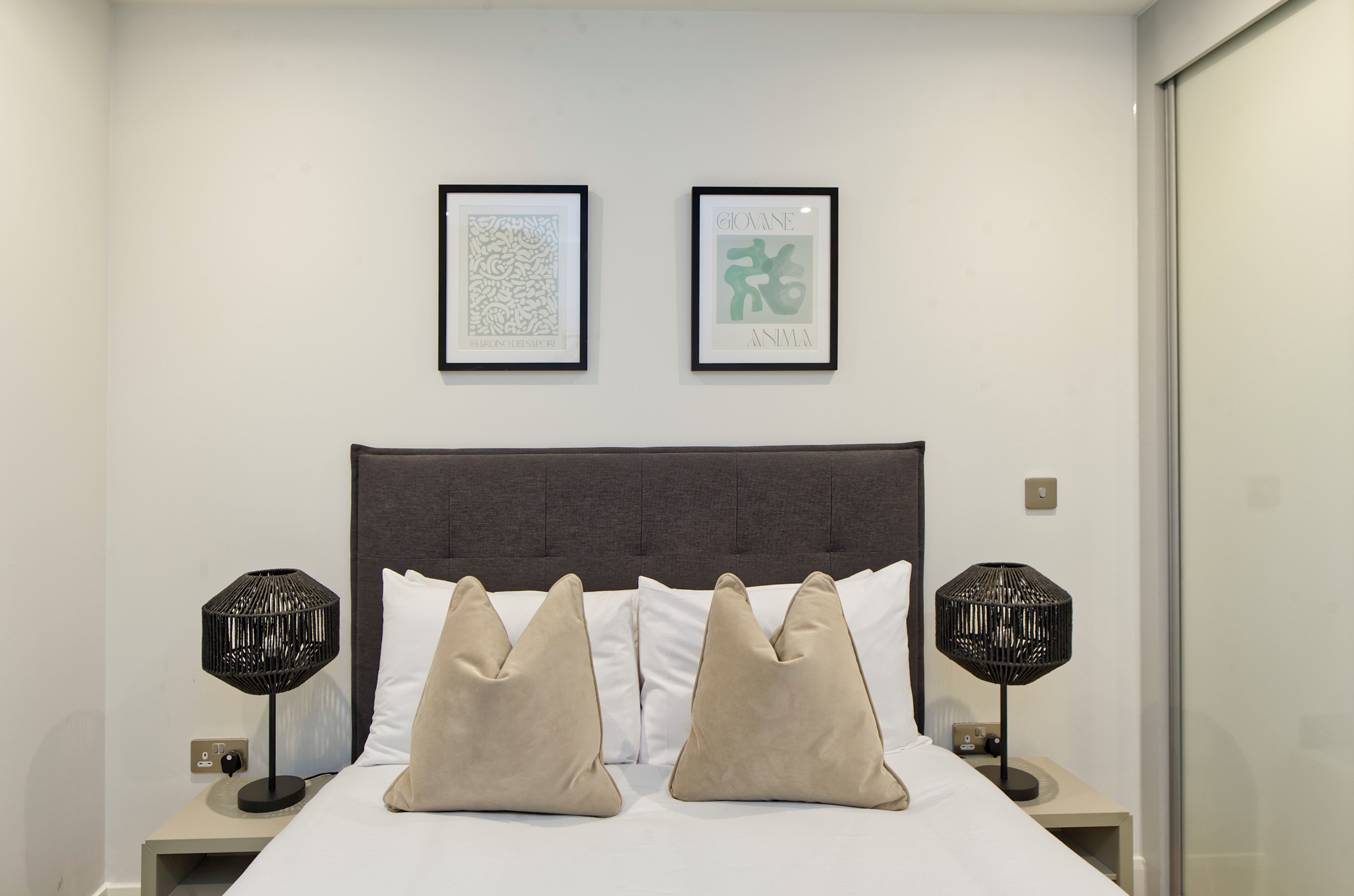Un lit bien fait, avec deux oreillers beige, une tête de lit foncée et deux lampes noires de chaque côté.