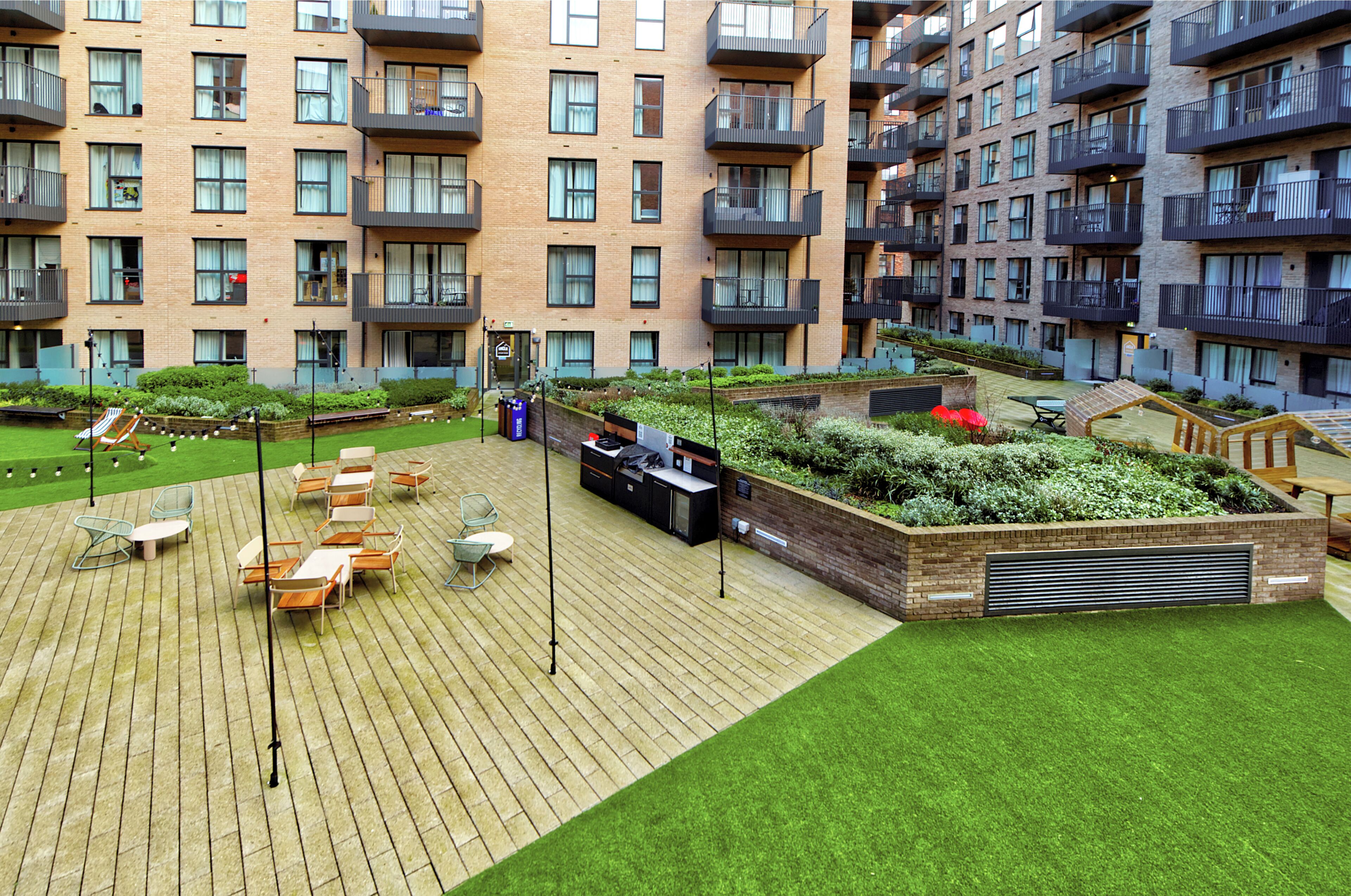 Un jardín en la azotea con mobiliario exterior, zona de barbacoa y vistas de un edificio residencial de varios pisos.