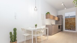 A modern kitchen with white cabinets, a built-in oven, and a refrigerator. There is a dining area with a white table and chairs, and a cactus plant on the floor. - GuestReady - Cozy stay near Malvarrosa Beach (Valencia)