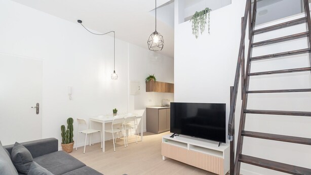 A modern living room with a grey sofa, a flat-screen TV on a white stand, a dining area with a white table and chairs, and a kitchen area with wooden cabinets and a hanging plant. - GuestReady - Cozy stay near Malvarrosa Beach (Valencia)