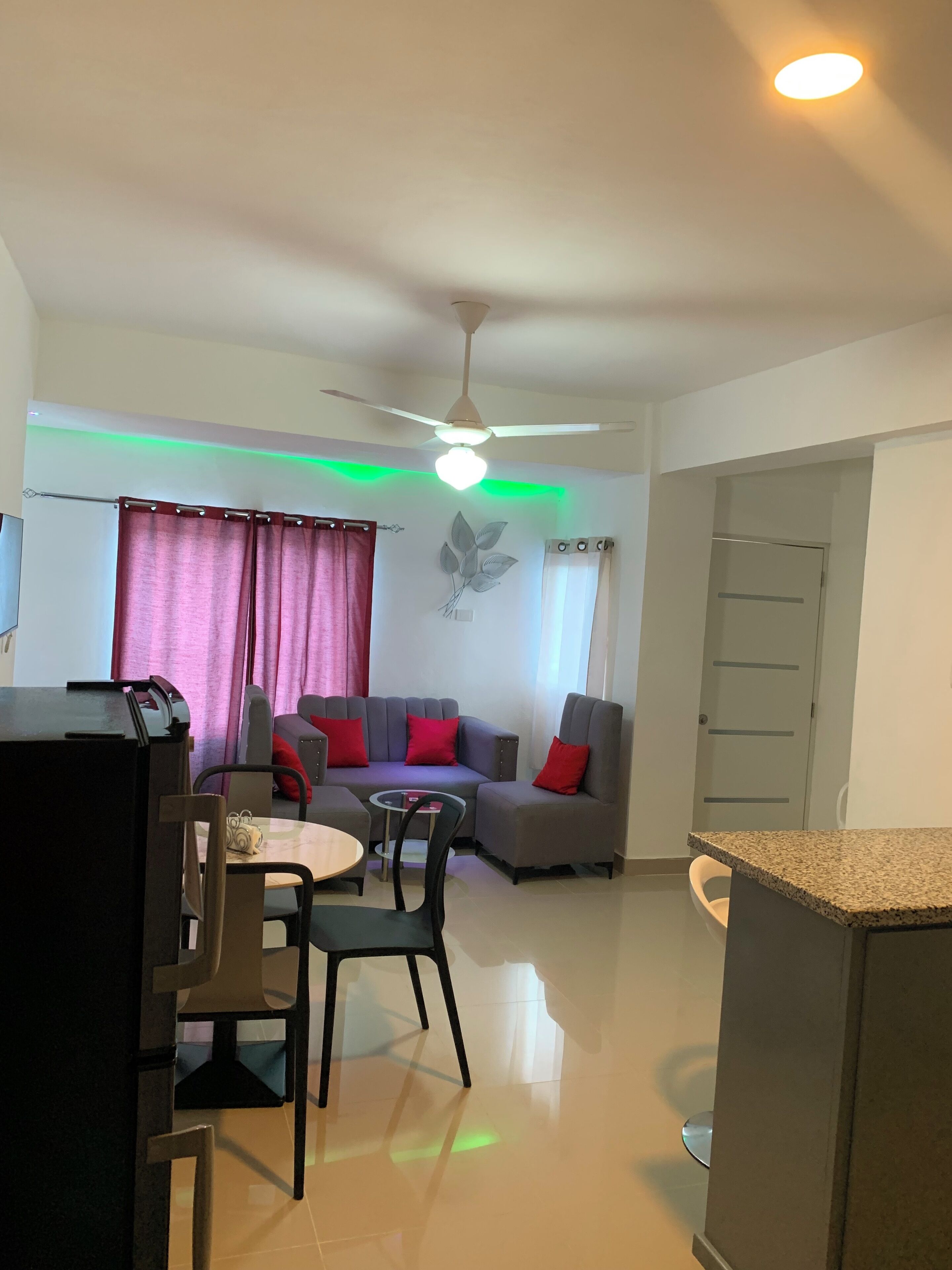 Een moderne woonkamer met een grijze bank, rode kussens, een eettafel met stoelen, een plafondventilator en een wandlamp.