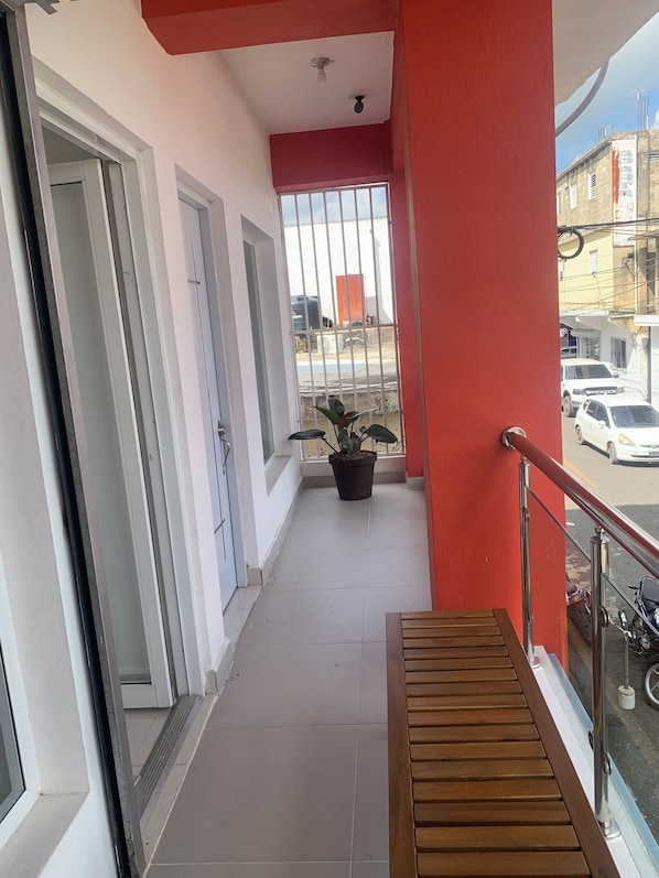 A balcony with a red wall, a potted plant, and a view of parked cars and buildings. - Plata View Apartahotel (Monte Plata)