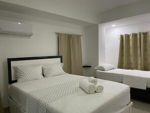 A hotel room with a single bed and a smaller bed, both with white linens and patterned bedspreads. There are two sets of beige curtains, one on each window. - Plata View Apartahotel (Monte Plata)