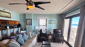A living room with a sofa, two armchairs, a coffee table, and a ceiling fan. There is a window with a view of the beach. - Ocean Breeze East 201 (Pensacola)