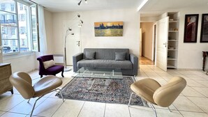 A modern living room with a grey sofa, two armchairs, a glass coffee table, a patterned rug, a floor lamp, and a painting on the wall.