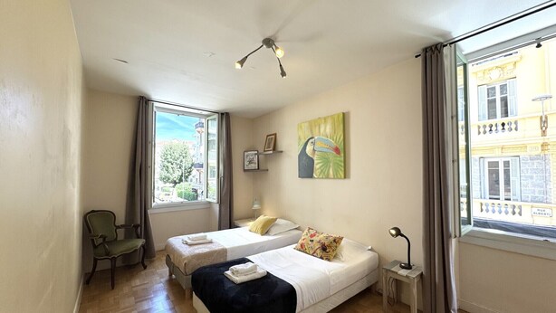 A hotel room with two beds, a chair, a window with curtains, a painting, and a lamp.