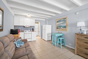 A compact living space with a kitchenette, a sofa with a turtle pillow, a small table with stools, and a painting on the wall. - Beachfront Studio | Amazing Sunsets | Pool (Panama City Beach)