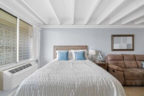 A hotel room with a bed, a reclining chair, a bedside table with a lamp, and a window with blinds. - Beachfront Studio | Amazing Sunsets | Pool (Panama City Beach)
