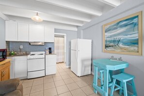 A kitchen with white cabinets, a stove, and a refrigerator. There is a blue table with two stools and a painting on the wall. - Beachfront Studio | Amazing Sunsets | Pool (Panama City Beach)