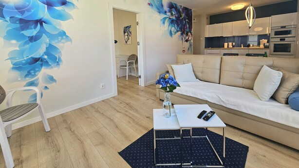 A modern living room with a sofa, a coffee table, and a vase of flowers. There is a kitchen area with cabinets and a refrigerator.