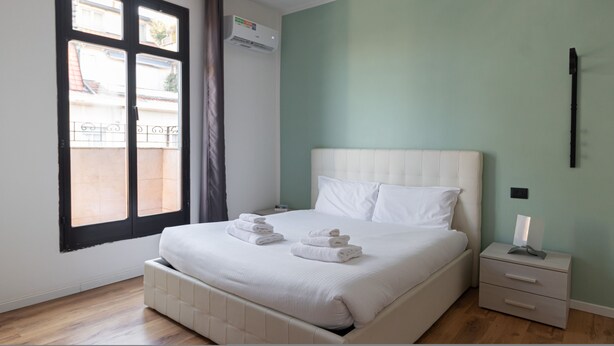 A neatly made bed with white linens and towels, a bedside table with a lamp, and a window with a view of buildings outside. - Italianway - Rosmini 11 B (Milan)