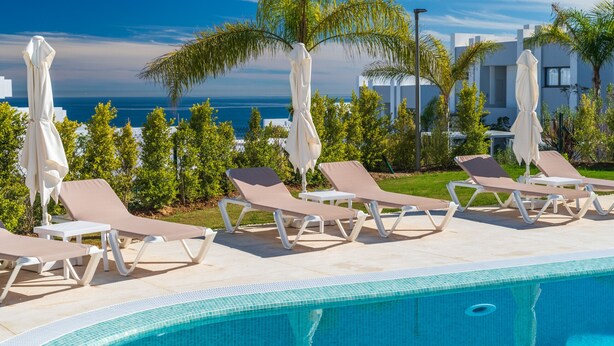 A poolside area with lounge chairs and umbrellas, a clear view of the sea, and a modern building in the background.