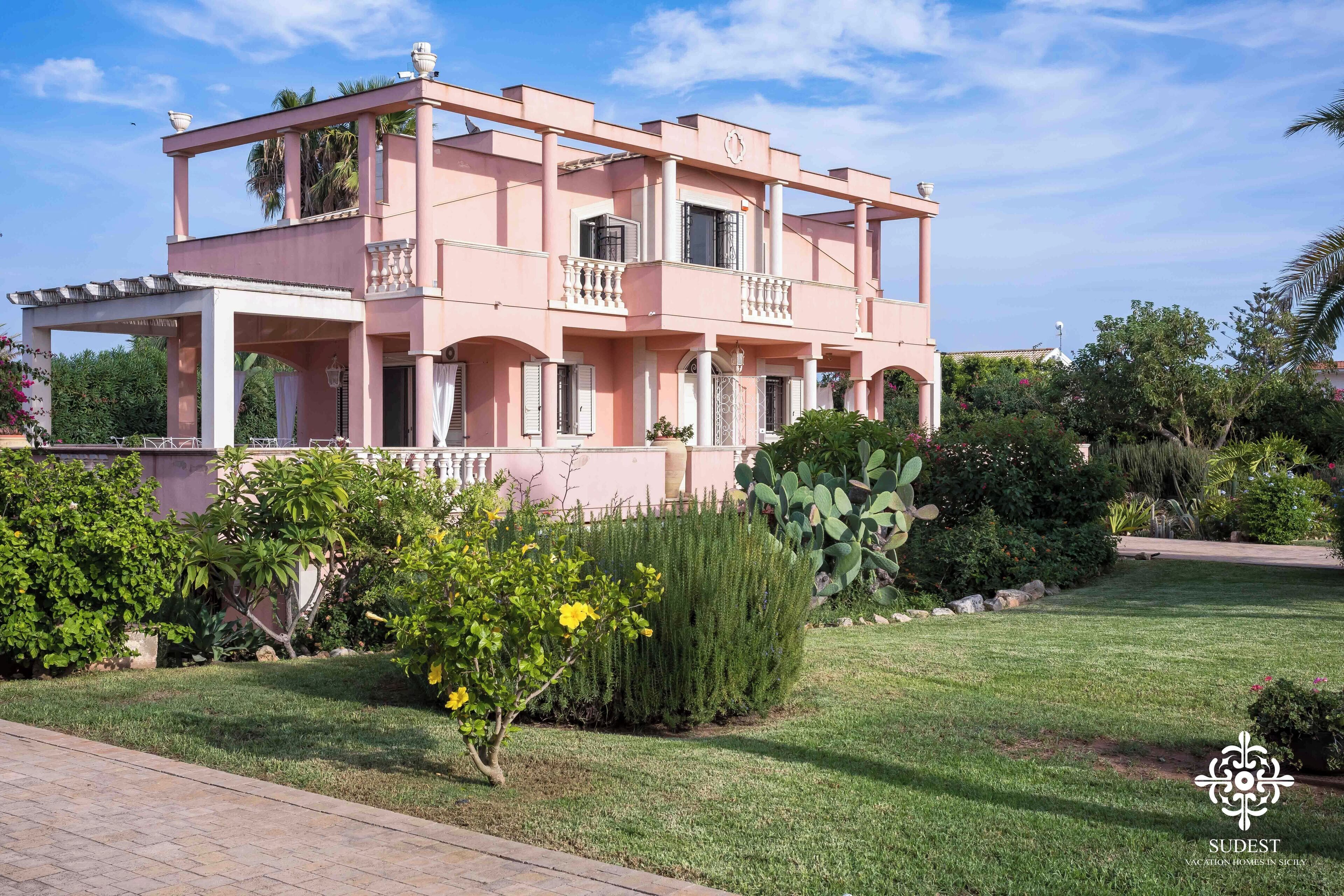 Een roze villa met balkon, omgeven door verzorgde tuinen en een heldere lucht.