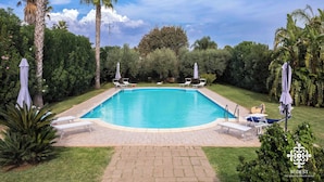 A rectangular swimming pool with a paved deck, surrounded by palm trees and greenery. - Villa Festosa - Luxury Retreat  (Santa Croce Camerina)