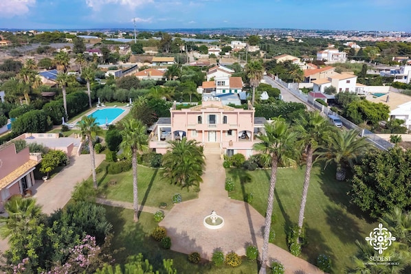An aerial view of a residential area with a prominent pink villa featuring a swimming pool, surrounded by lush greenery and palm trees.