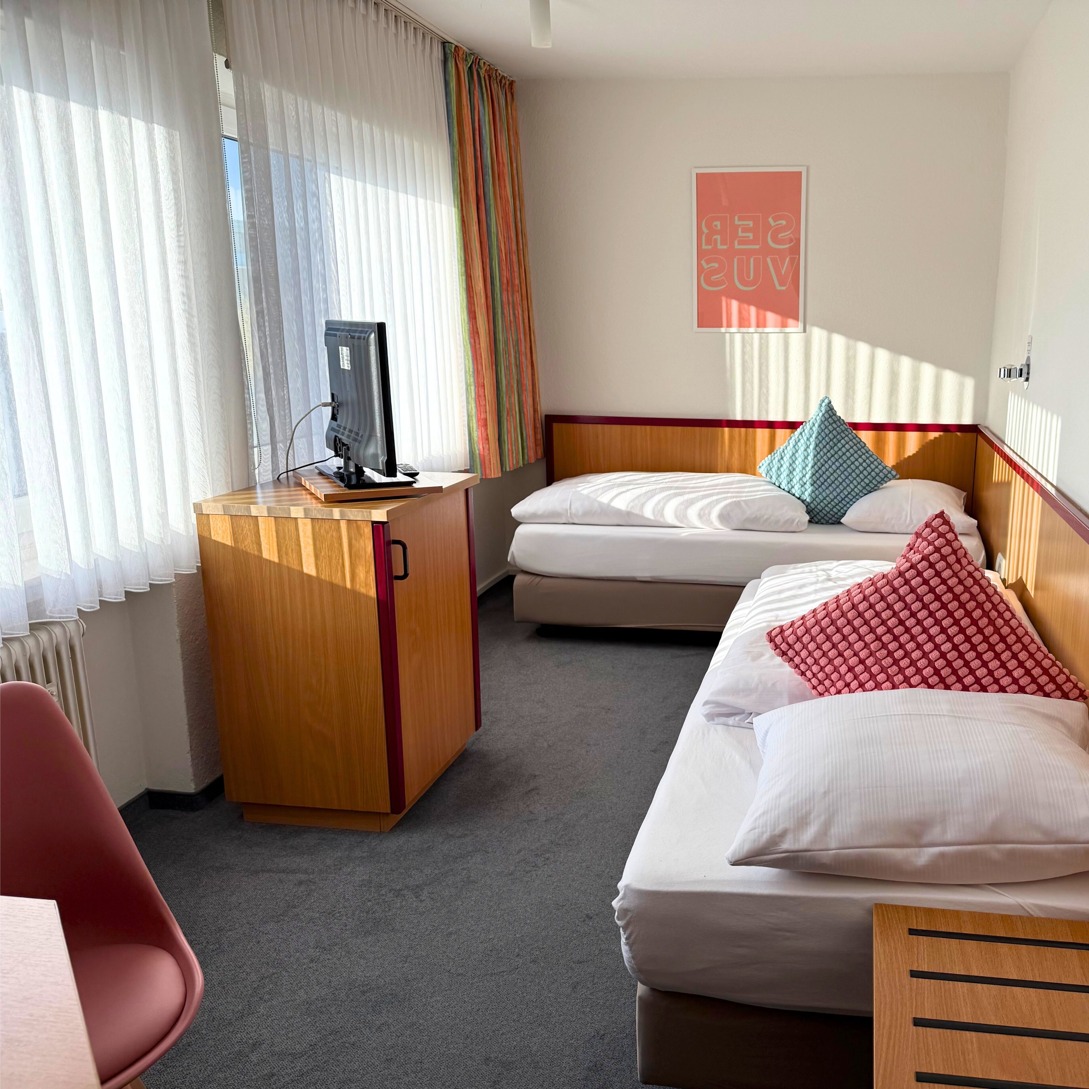 A hotel room with a wooden desk, a computer, a bed with pillows, and a poster on the wall.