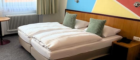 A hotel room with a bed, a bedside table, a nightstand, a radiator, and a wall with a colorful geometric design.