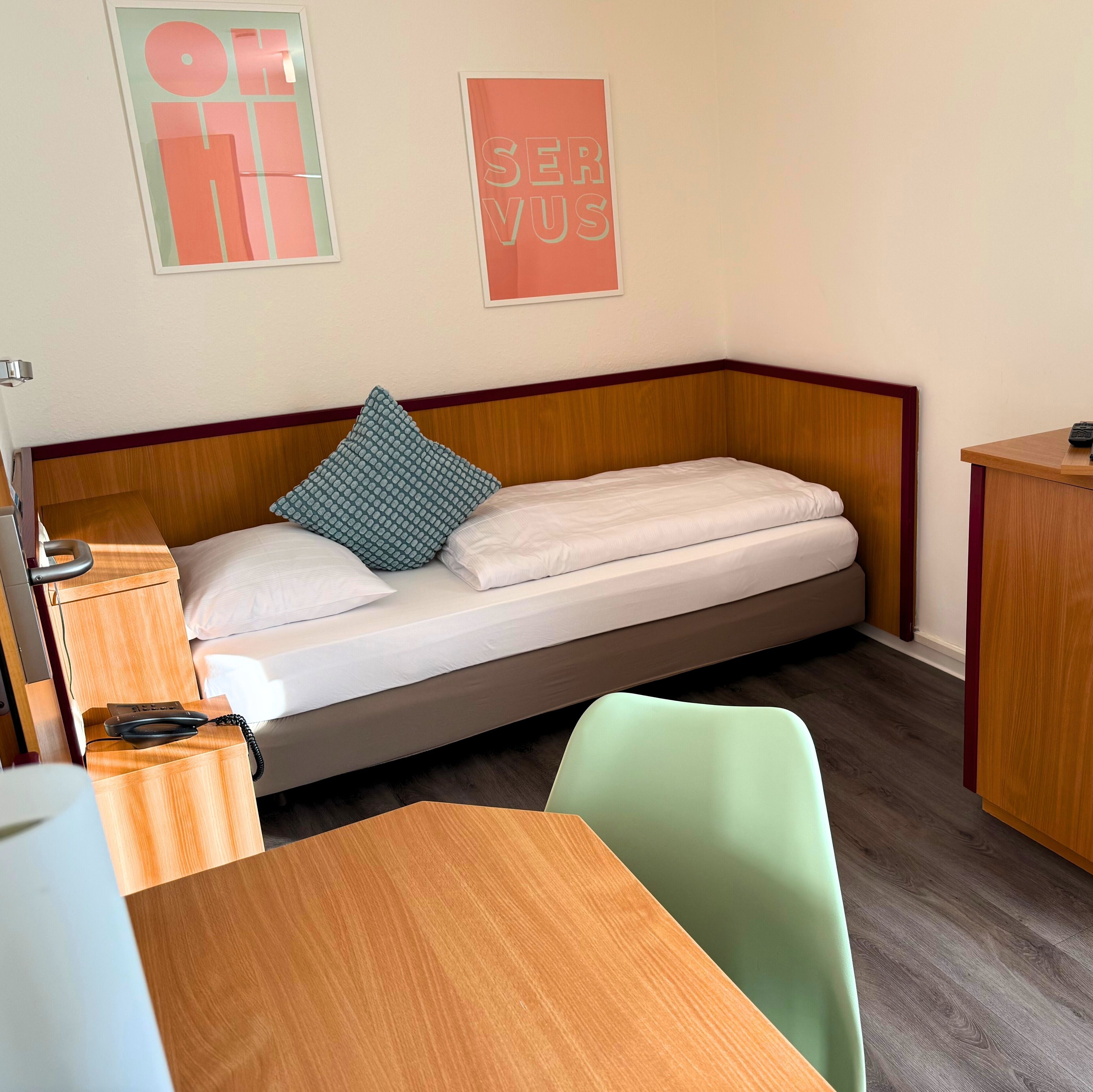 Een enkel bed met een houten hoofdbord, een bureau met telefoon, een stoel en een klein tafeltje.