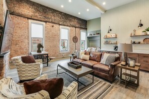 Ein geräumiges Wohnzimmer mit Backsteinwand, Ledersofas, einem Couchtisch und einer Küchenzeile mit Regalen und einer Uhr.