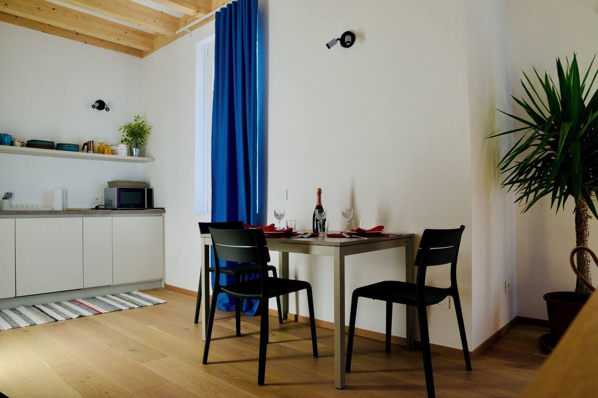 Ein Essbereich mit einem Holztisch, zwei Stühlen, einem blauen Vorhang, einer Topfpflanze und einer Mikrowelle auf der Arbeitsplatte.