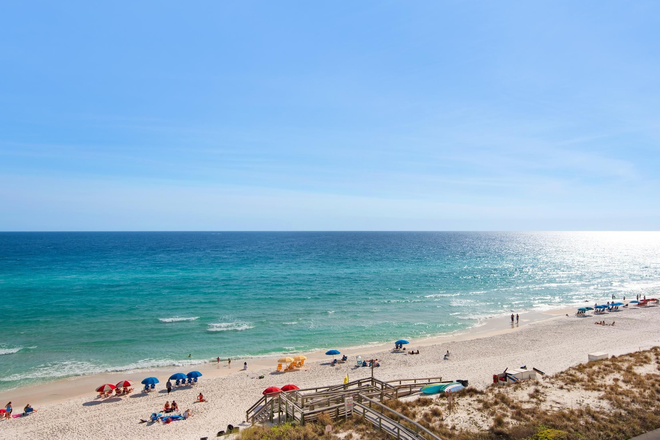 A beach with a wooden walkway leading to the water, umbrellas, and people relaxing.