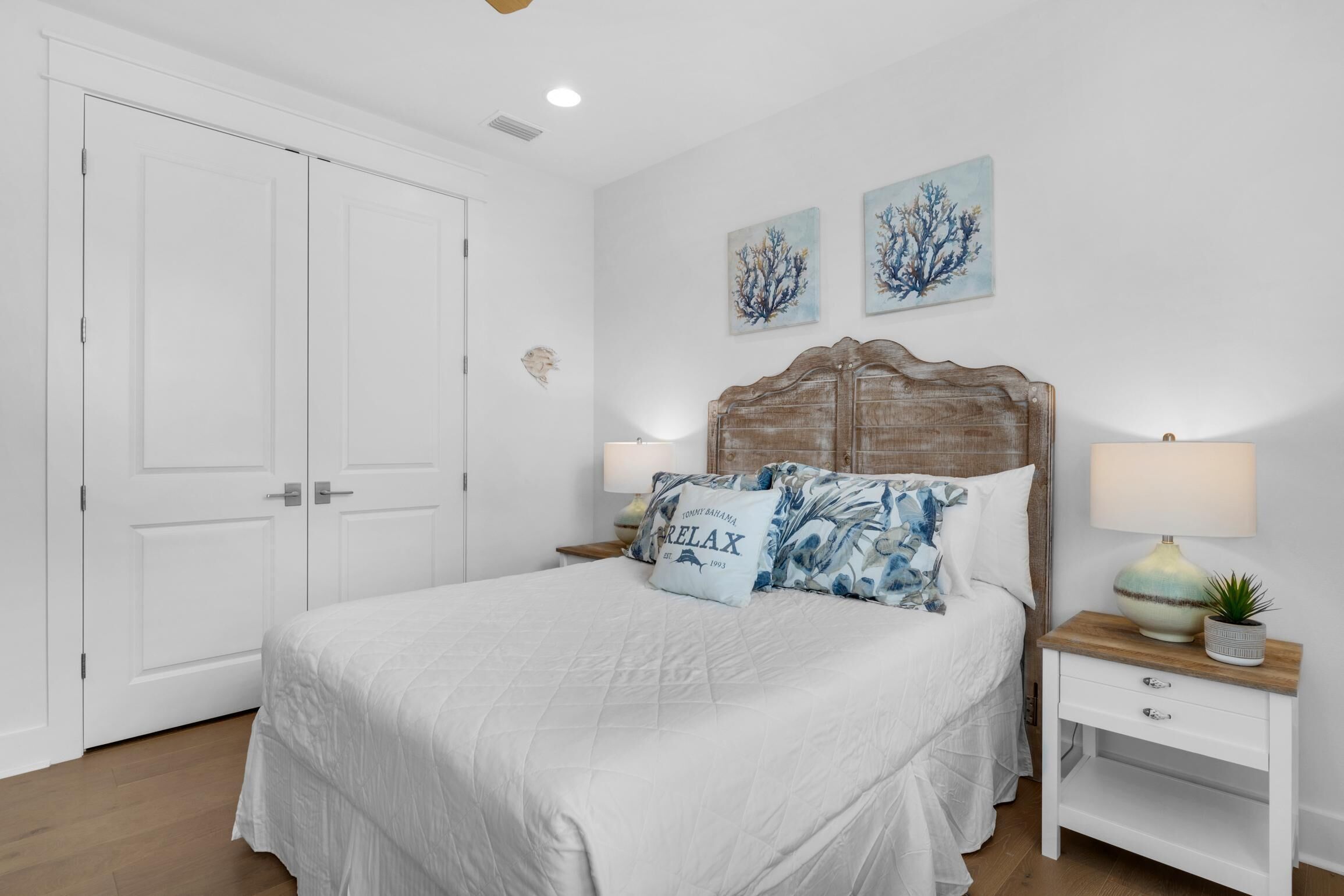 Ein Schlafzimmer mit einem großen Bett, weißen Wänden und einem hölzernen Kopfteil. Es sind zwei Wandbilder, ein Nachttisch mit einer Lampe und ein kleiner Topfpflanze zu sehen.