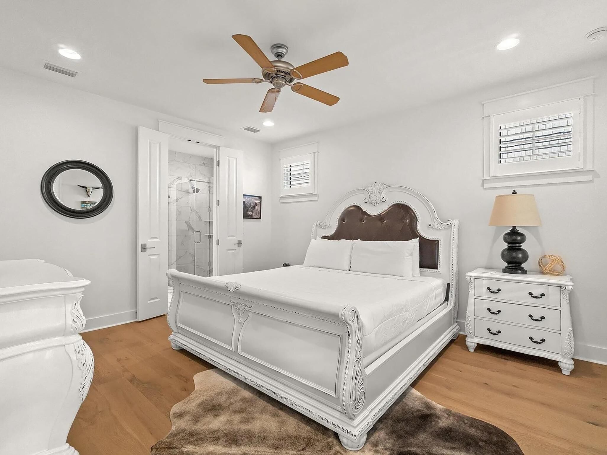 A bedroom with a white bed, a ceiling fan, a nightstand with a lamp, and a mirror on the wall.