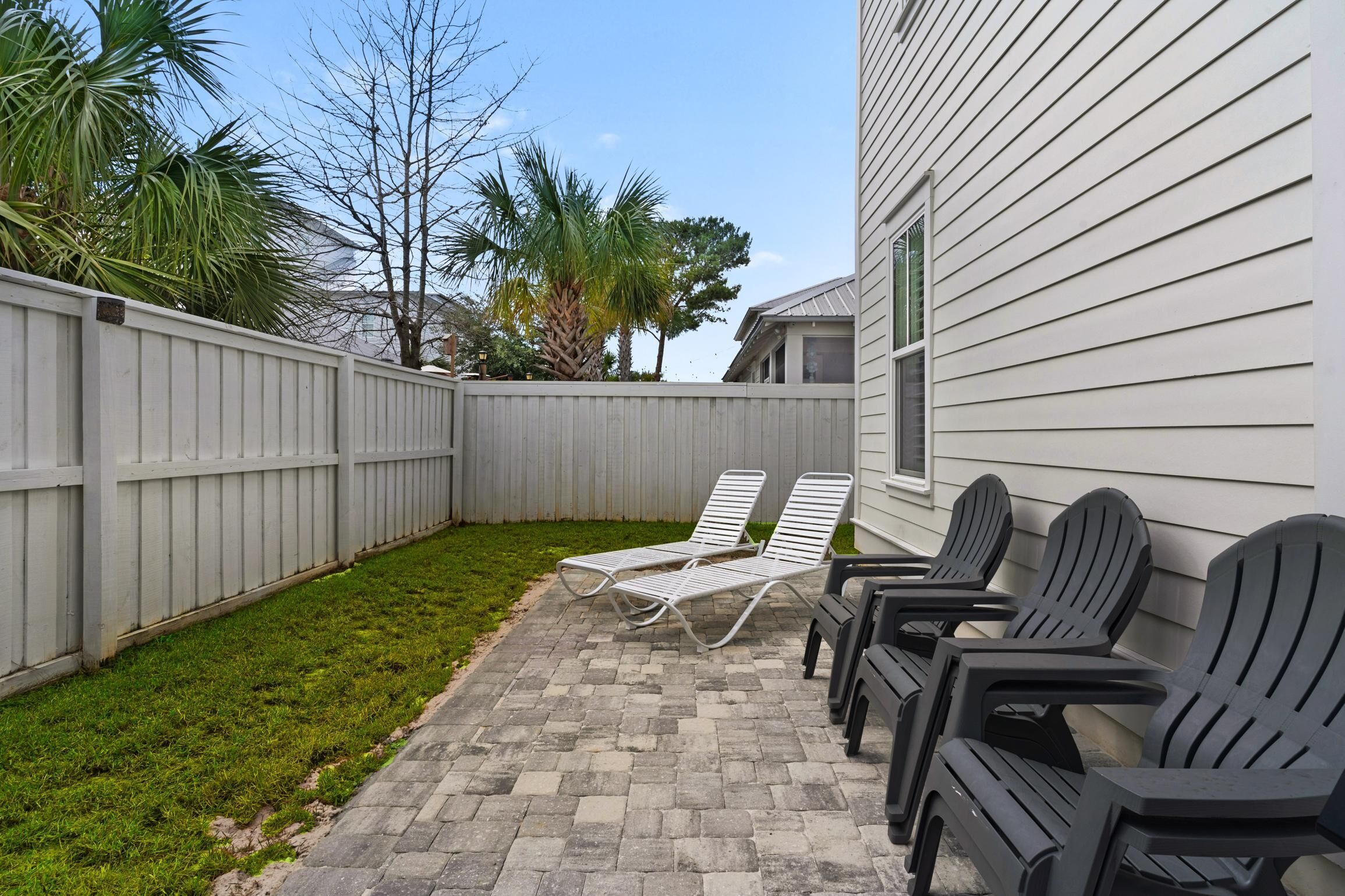A backyard with a paved patio, two white lounge chairs, and a row of black Adirondack chairs.